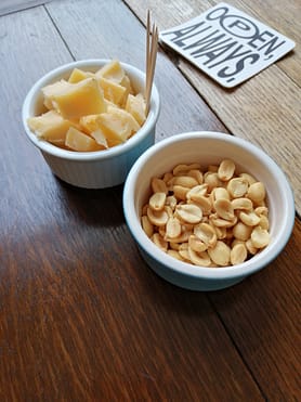 Two ramekins on a wooden table; nearground one filled salted peanuts, further one with cubes of cheese & cocktail sticks