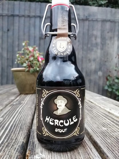 Dark brown flip top stubby bottle on the slat of a wooden picnic bench, label depicts Hercule Poirot.