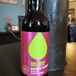 Bottle sits on a dark brown surface with a dark brown curved pillar beside it. Background is a blue wall with a few blurry items. Bottle has a shiny pink label with large yellowish-green teardrop shape on it