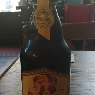 Dark brown stubby bottle with flip-top lid on a wooden table; yellow label depicts a stylised warrior with shield and spear