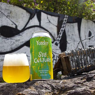 A green can sits beside a glass of blonde beer with a frothy head. Behind is a graffiti'd wall