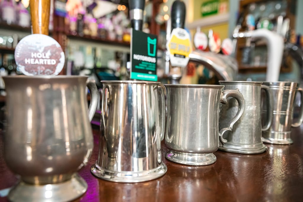 Five different shaped pewter tankards are arranged on a wooden bar top. They are getting further away from left to right of the image