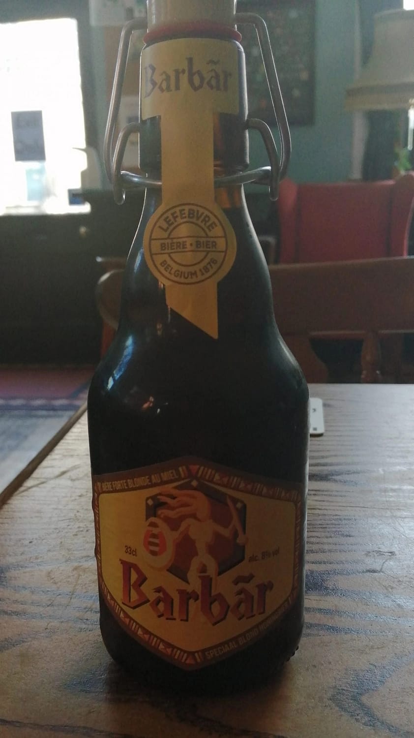 Dark brown stubby bottle with flip-top lid on a wooden table; yellow label depicts a stylised warrior with shield and spear