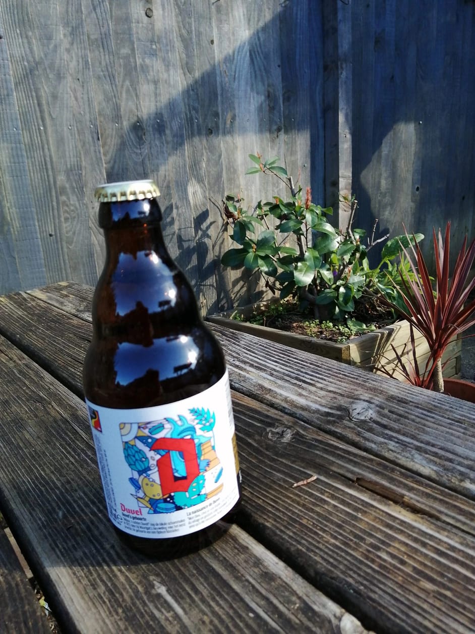 Stubby brown bottle on a picnic bench with a fence and plants in the background